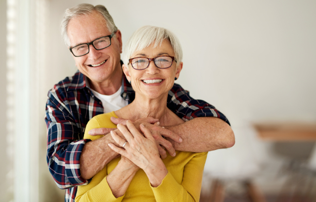 Senior couple smiling together in home setting, wearing glasses and casual clothing.