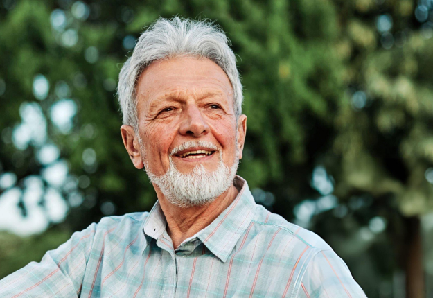 Smiling older man with gray hair and beard outdoors in front of green trees.