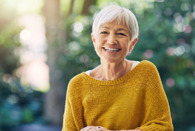 Smiling senior woman wearing yellow sweater outdoors in natural sunlight and greenery.
