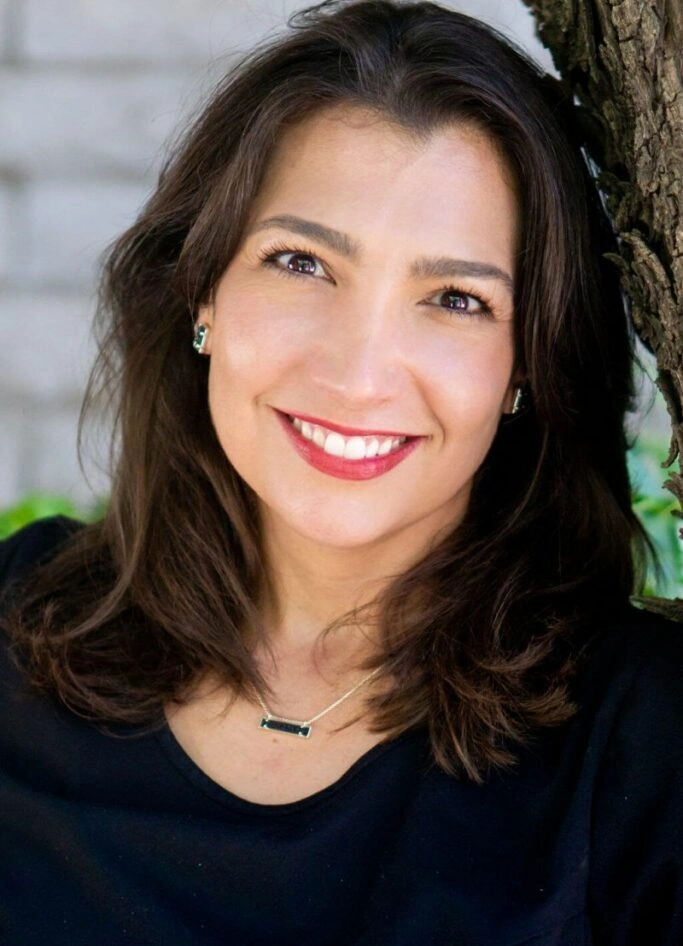 Professional headshot of smiling woman with dark hair against blurred outdoor background.
