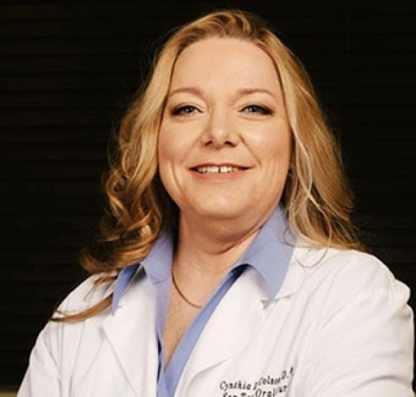 Professional headshot of female dentist in white coat and blue scrubs against dark background.